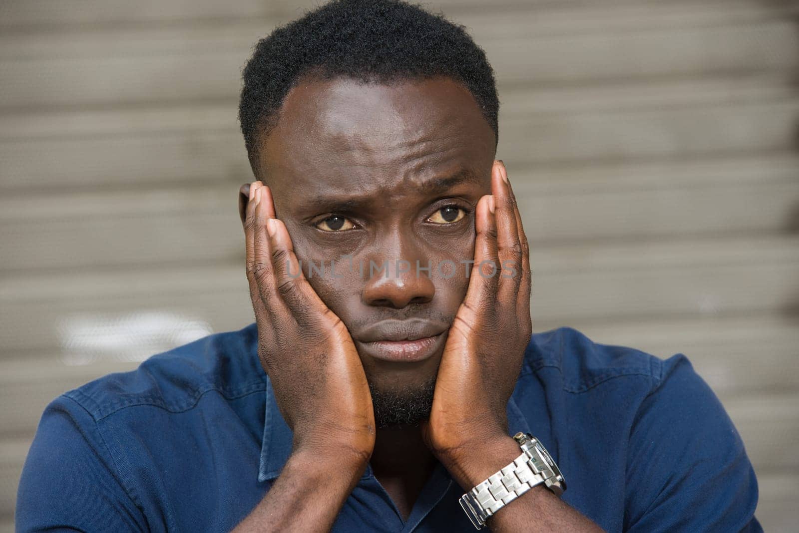 young man in blue shirt sitting outdoors thinking of hands on cheeks.