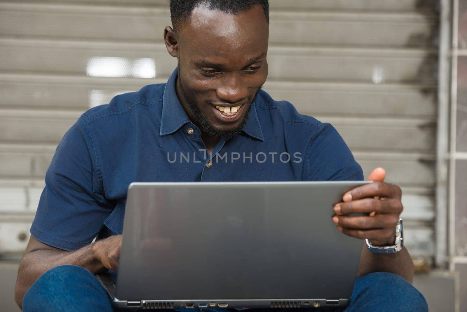 young man in blue shirt sitting outdoors looking at laptop smiling.