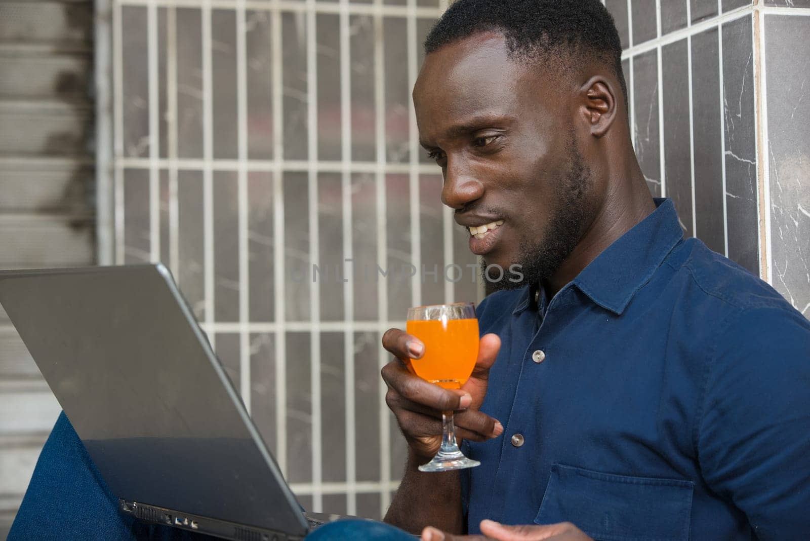 young man in blue shirt sitting near a wall looking at laptop smiling with a glass of juice in hand.