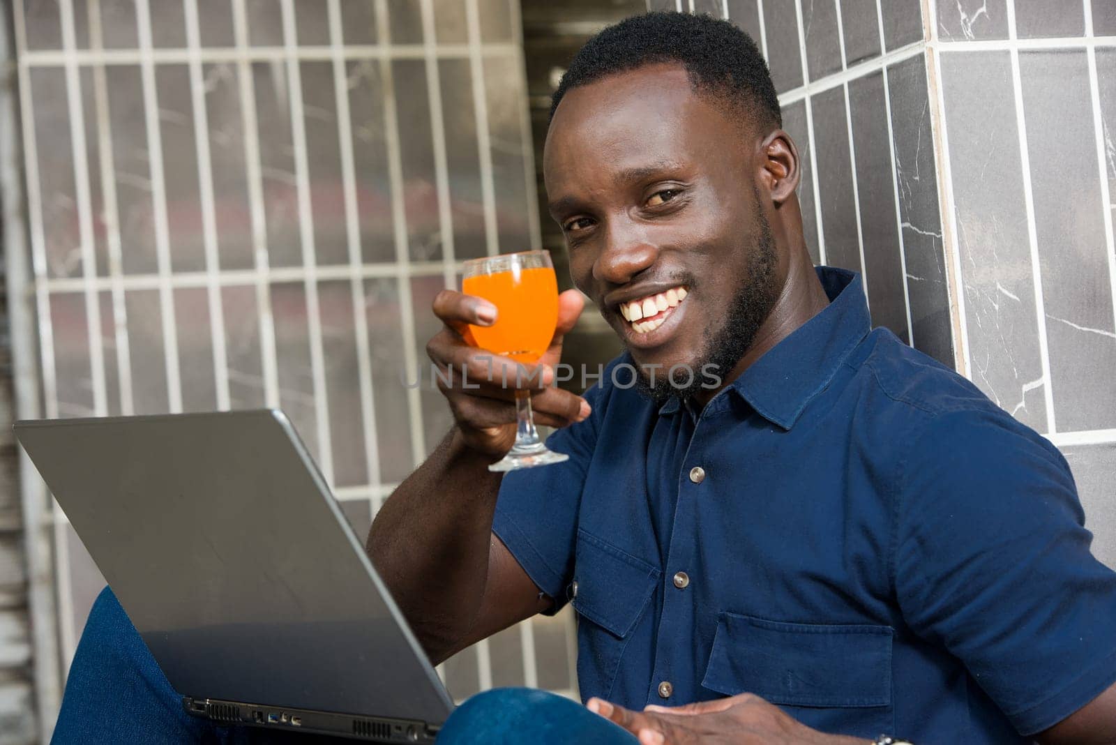 young man in blue shirt sitting near a wall looking at laptop smiling with a glass of juice in hand.