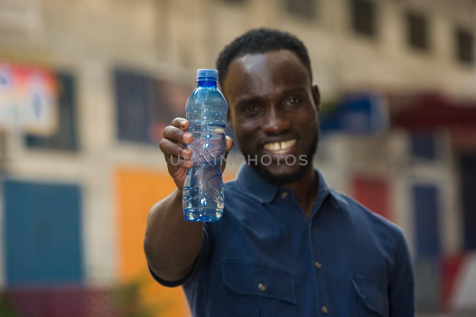 young man standing outdoors presenting a bottle of water while smiling.