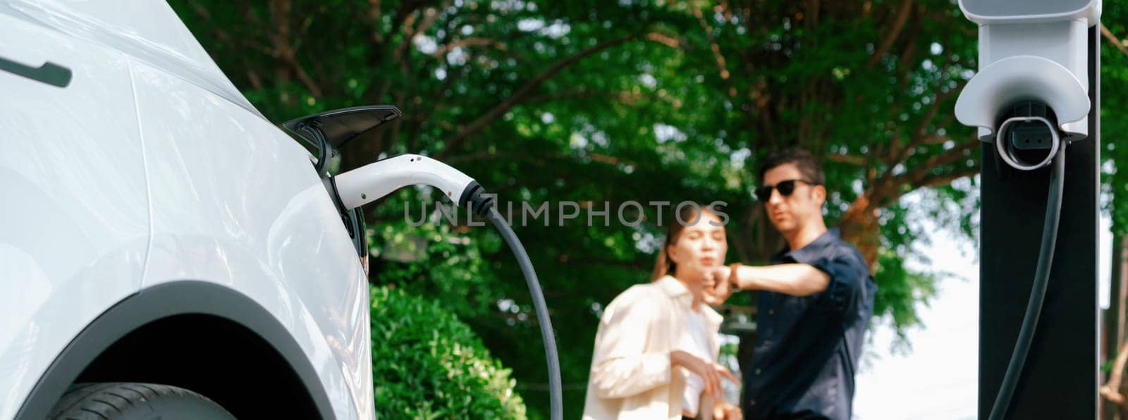 Young couple recharge electric car battery from charging station in green city park in springtime. Rechargeable EV car for sustainable environmental friendly urban travel lifestyle. Panorama Expedient