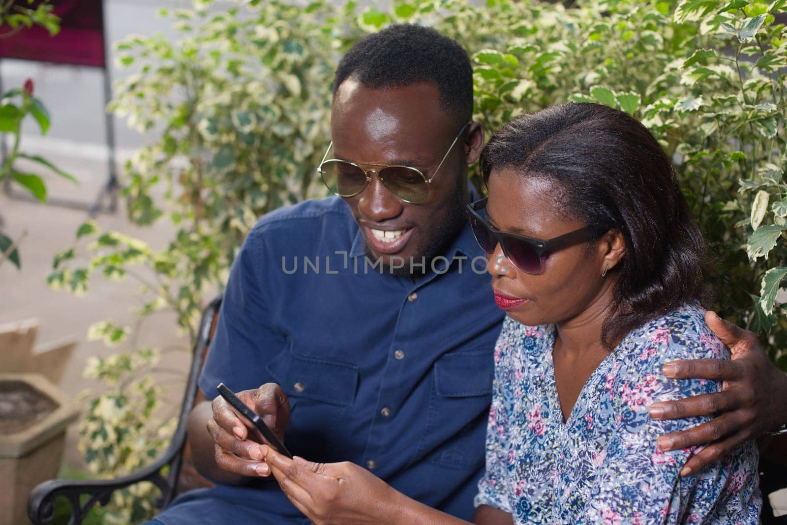 young couple sitting in a park looking at mobile phone while smiling.