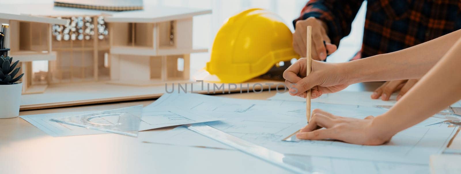 Cropped image of cooperative architect team decide and work together on meeting table with house model, safety helmet and architectural plan scatter around. Closeup. Focus on hand. Burgeoning.