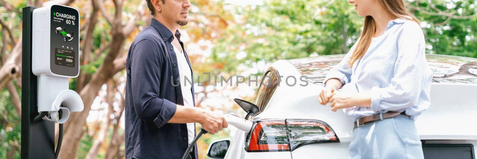 Panorama banner of lovely young couple recharging battery for electric car during autumnal road trip travel EV car in autumnal forest. Eco friendly travel on vacation during autumn. Exalt