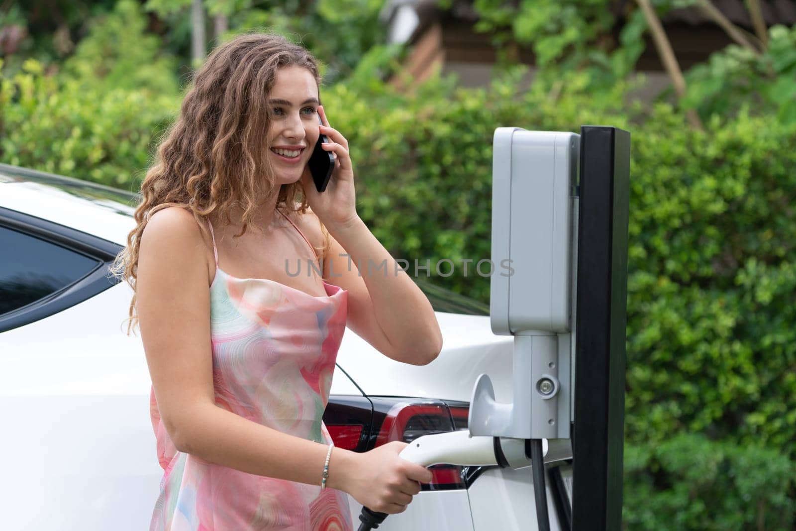 Modern eco-friendly woman recharging electric vehicle from home EV charging station. Innovative EV technology utilization for tracking energy usage to optimize battery charging at home. Synchronos