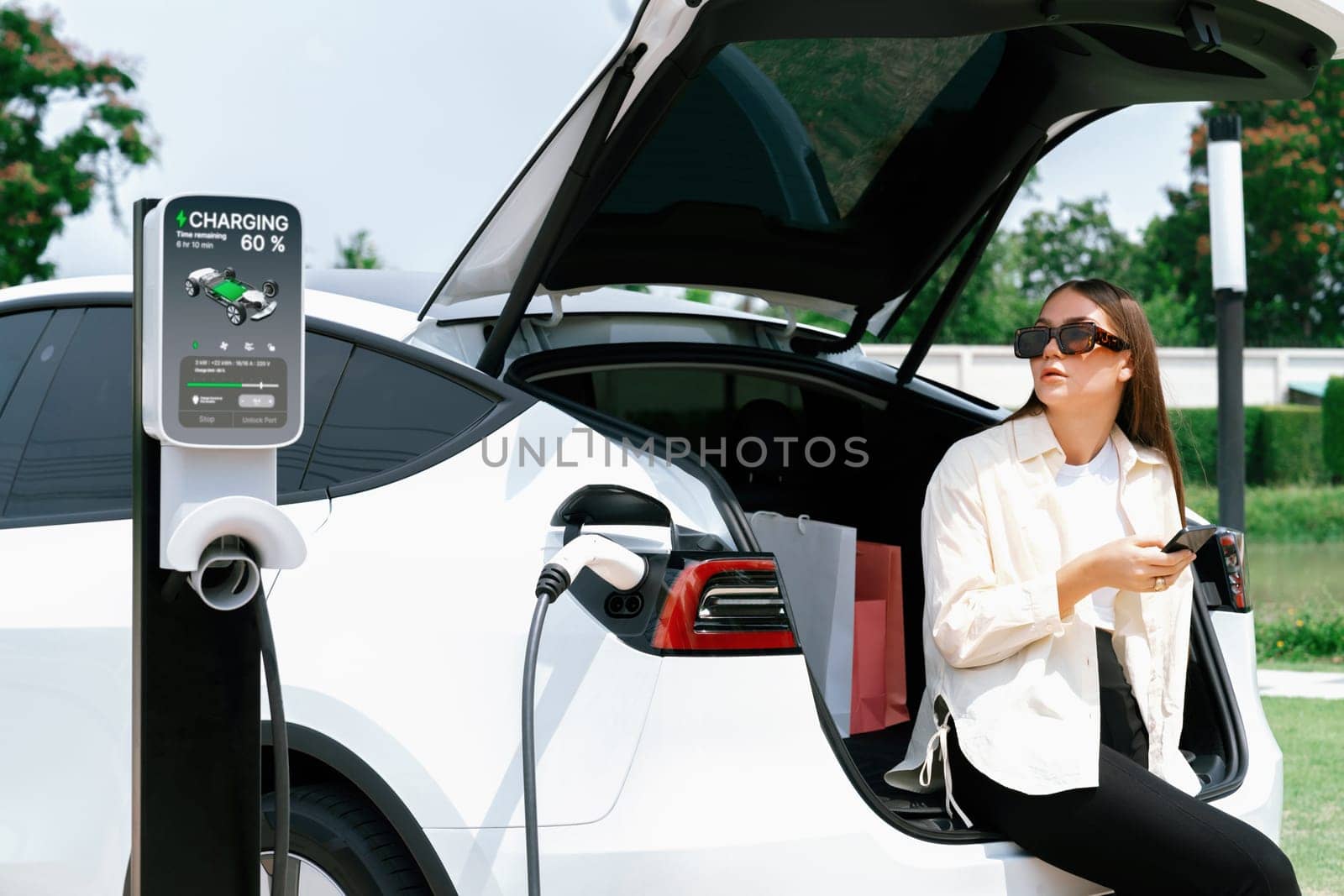 Young woman use smartphone to pay for electricity at public EV car charging station green city park. Modern environmental and sustainable urban lifestyle with EV vehicle. Expedient