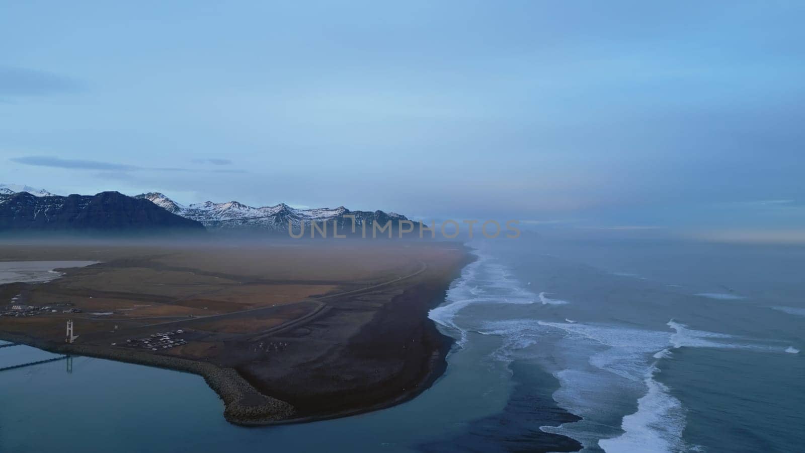 Aerial view of icelandic coastline with famous black sand beach and nordic scenery, snowy mountains and hills. Beautiful altantic ocean shore with roadside, natural panoramic view. Slow motion.