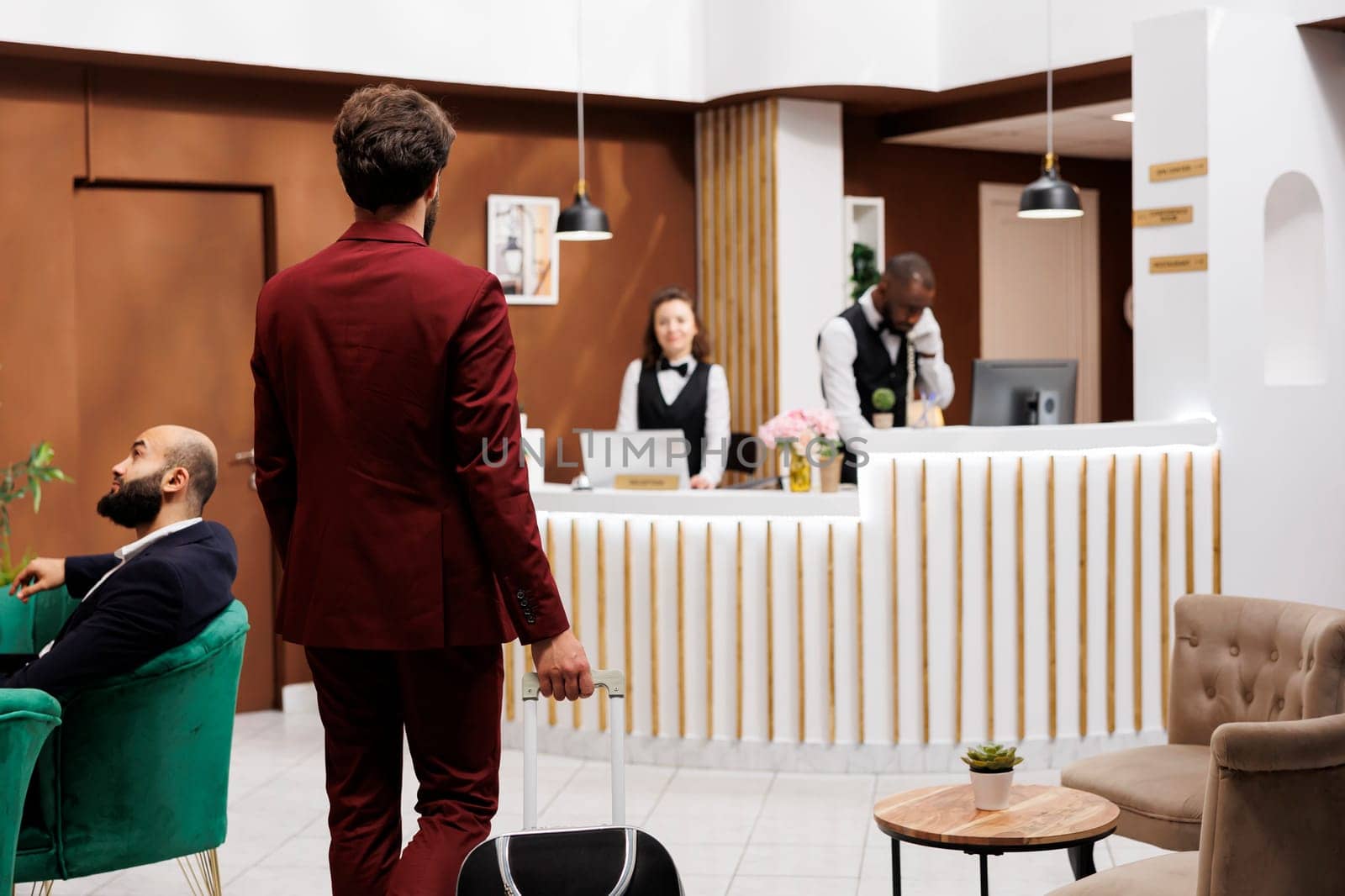 Formal guest enter reception area, carrying suitcase at front desk to do check in process. Businessman in suit with luggage trolley travelling for work purposes, staff ensuring pleasant stay.