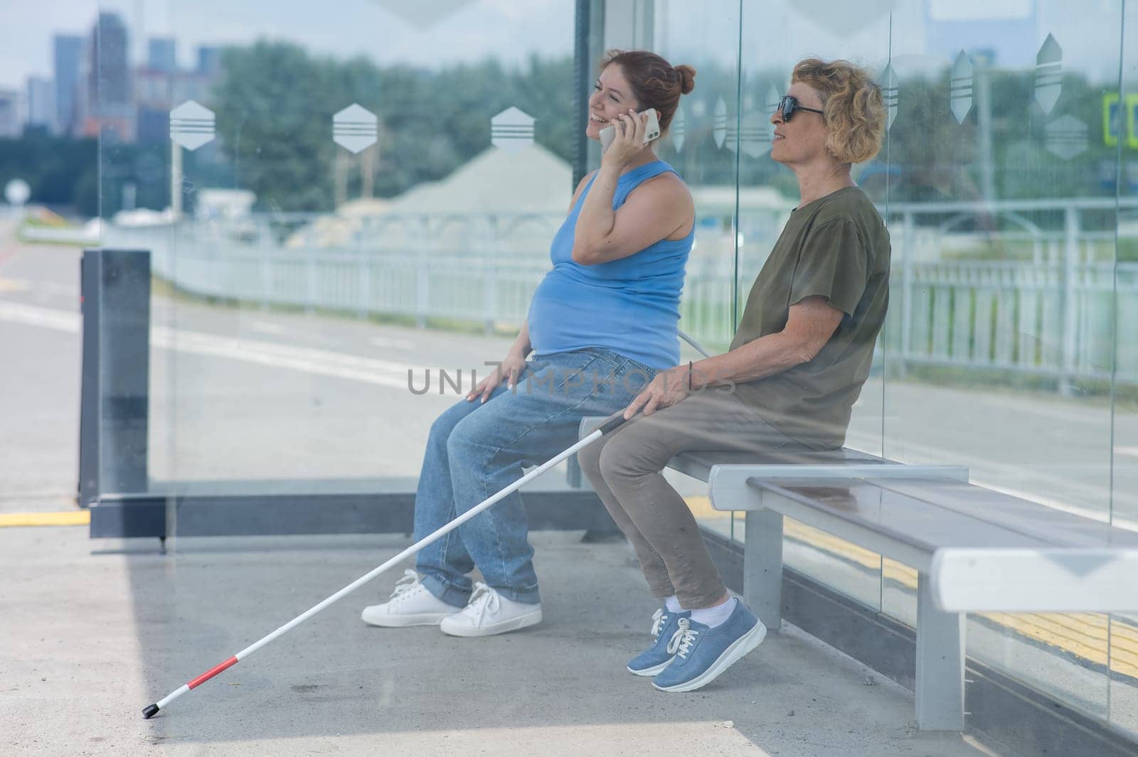 An elderly blind woman and a pregnant woman are sitting at a bus stop and waiting for the bus. by mrwed54
