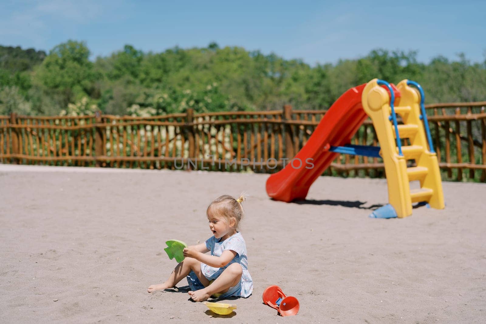 Little girl plays with plastic molds while sitting on the sand at the playground by Nadtochiy