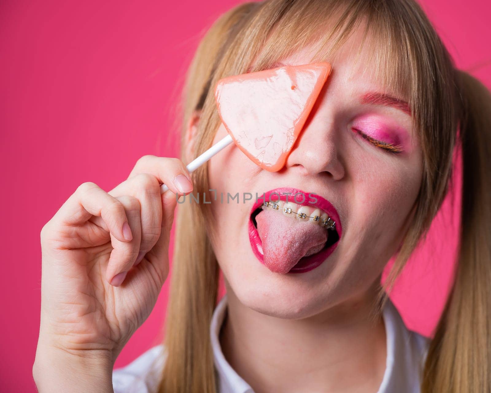 Portrait of a young woman with braces and bright makeup eating a lollipop on a pink background