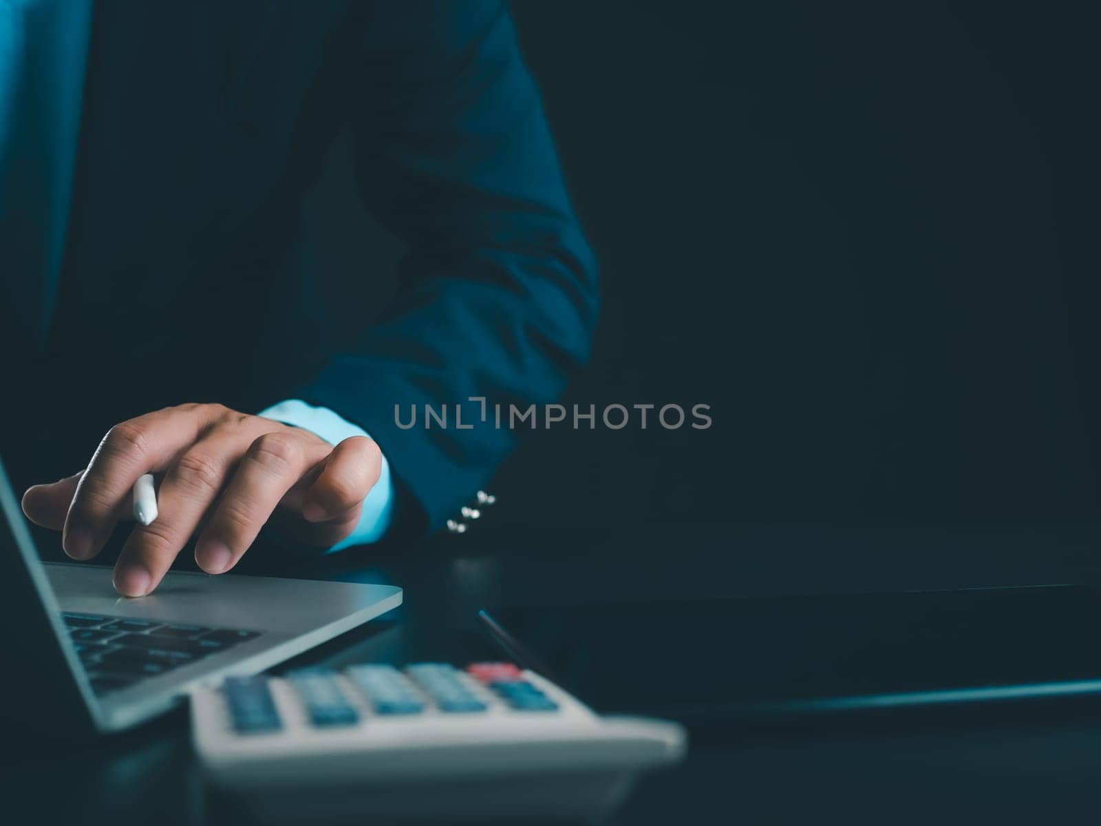 Businessman in black suit working on laptop computer and tablet and calculator, Hand Hands touch on tablet at office with dark background, Online working, Close up, Copy space.