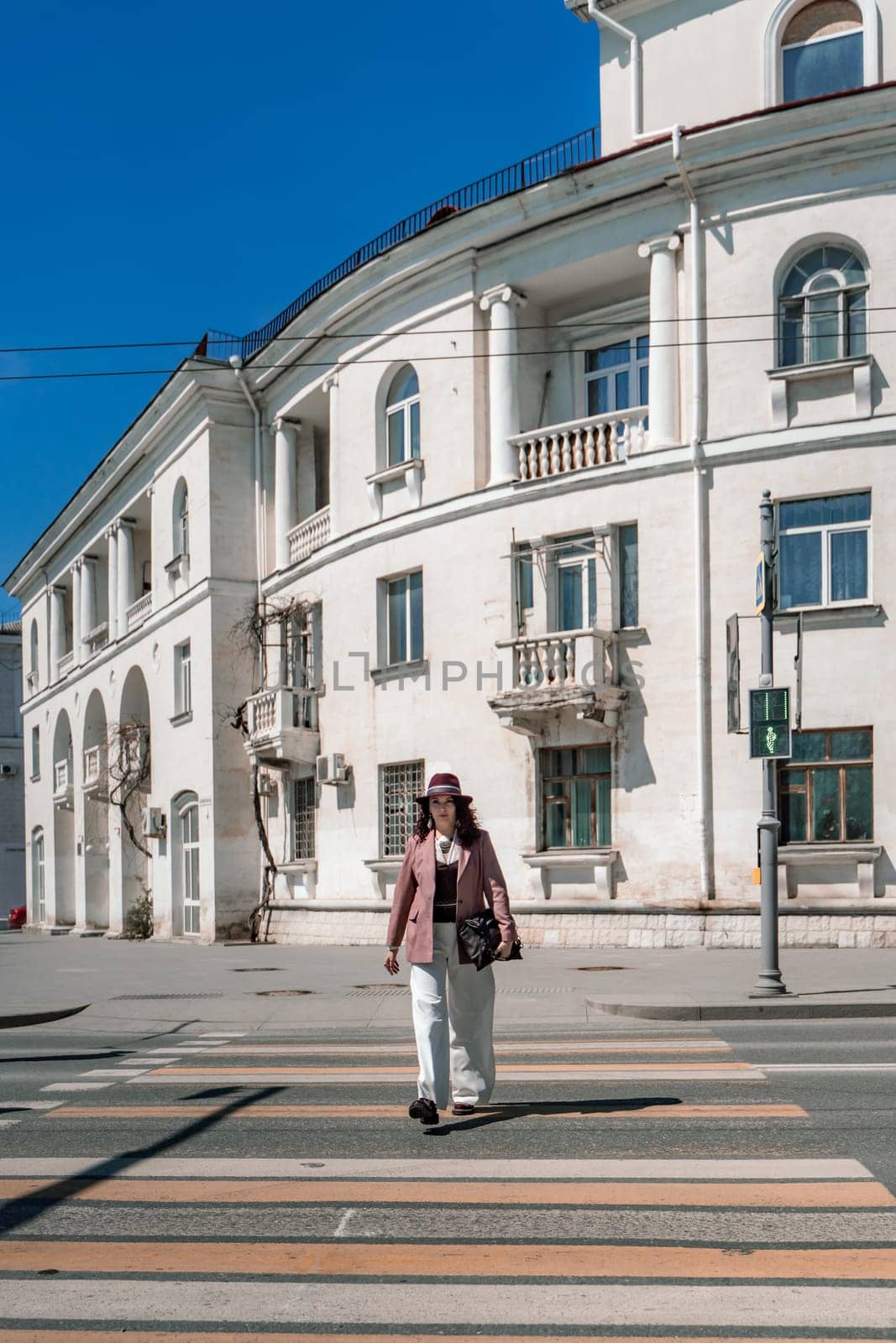 Woman city road crossing. Stylish woman in a hat crosses the road at a pedestrian crossing in the city. Dressed in white trousers and a jacket with a bag in her hands. by Matiunina