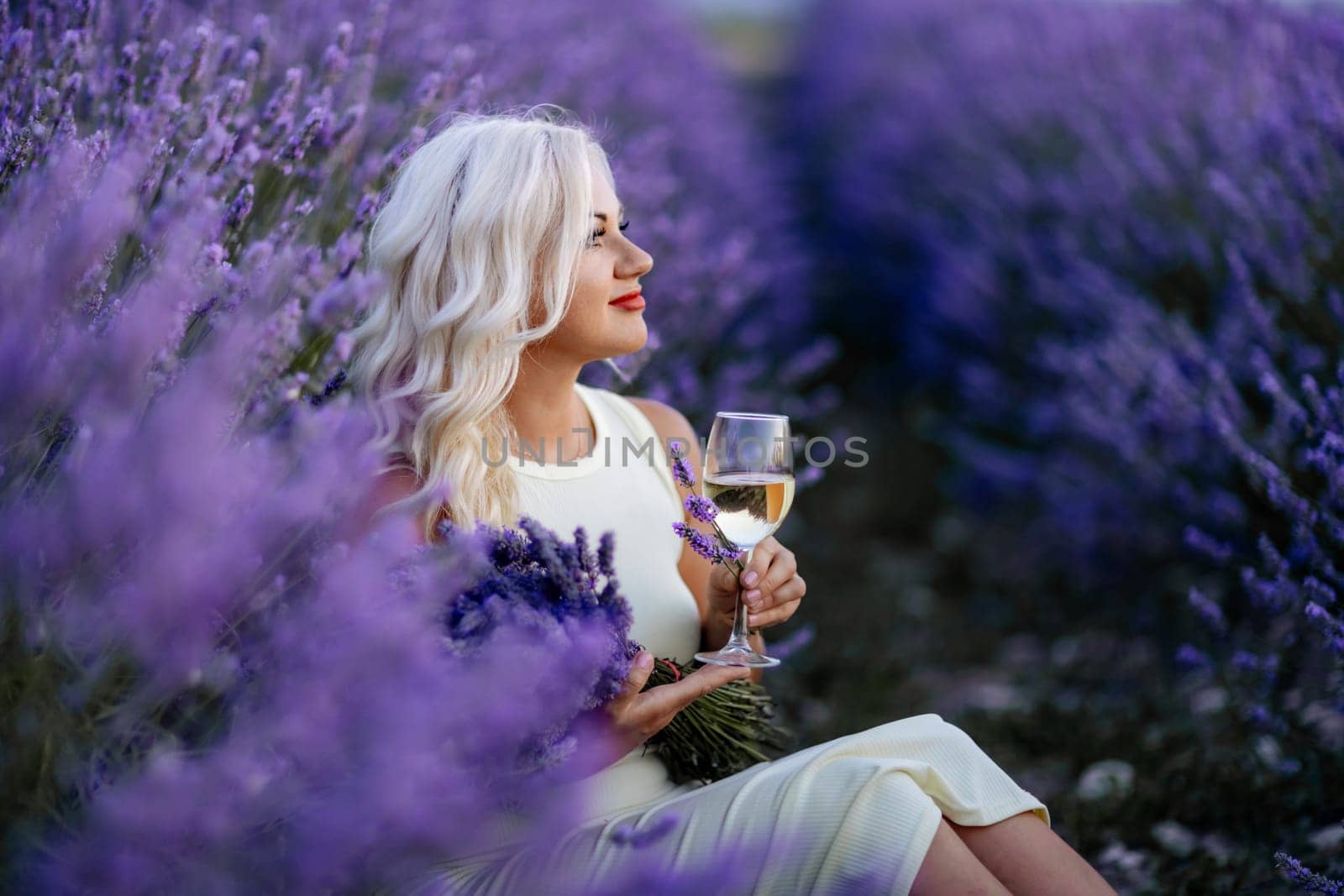 Blonde lavender field holds a glass of white wine in her hands. Happy woman in white dress enjoys lavender field picnic holding a large bouquet of lavender in her hands . Illustrating woman's picnic in a lavender field