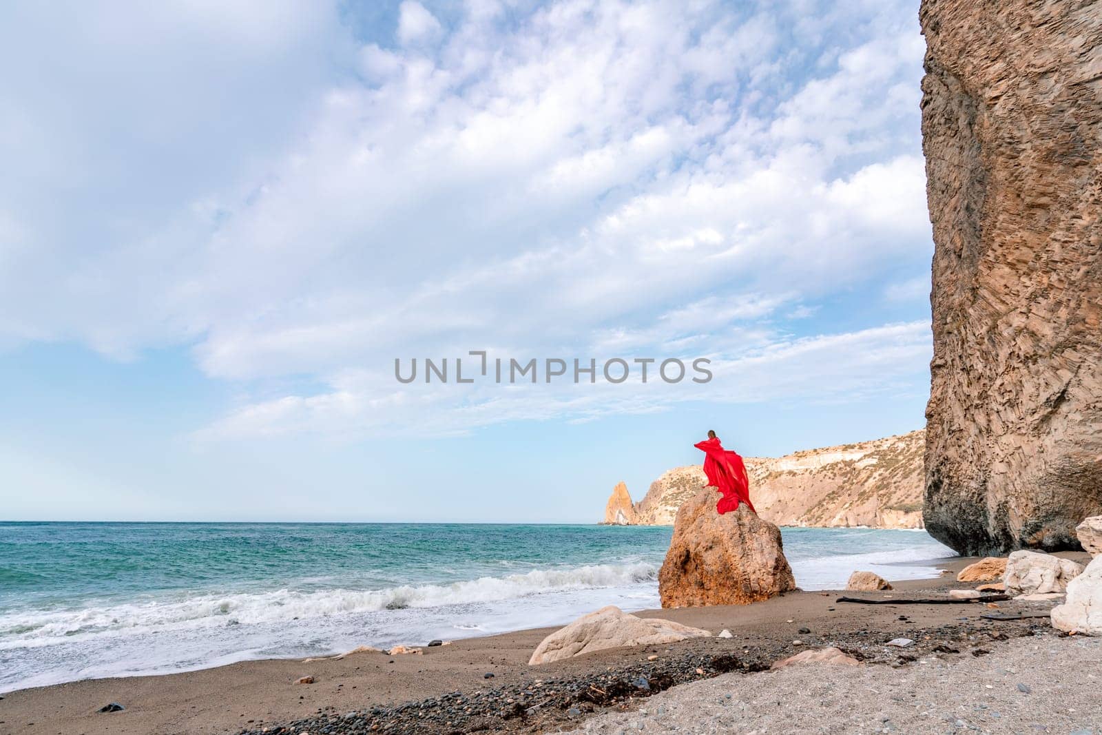 woman sea red dress. Woman with long hair on a sunny seashore in a red flowing dress, back view, silk fabric waving in the wind. Against the backdrop of the blue sky and mountains on the seashore