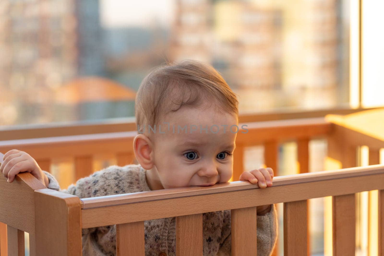 a little girl sadly gnaws the edge of her crib with her teeth while waiting for her parents.