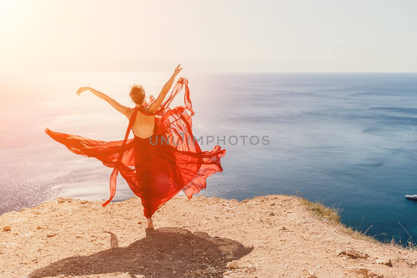 Side view a Young beautiful sensual woman in a red long dress posing on a rock high above the sea during sunrise. Girl on the nature on blue sky background. Fashion photo.