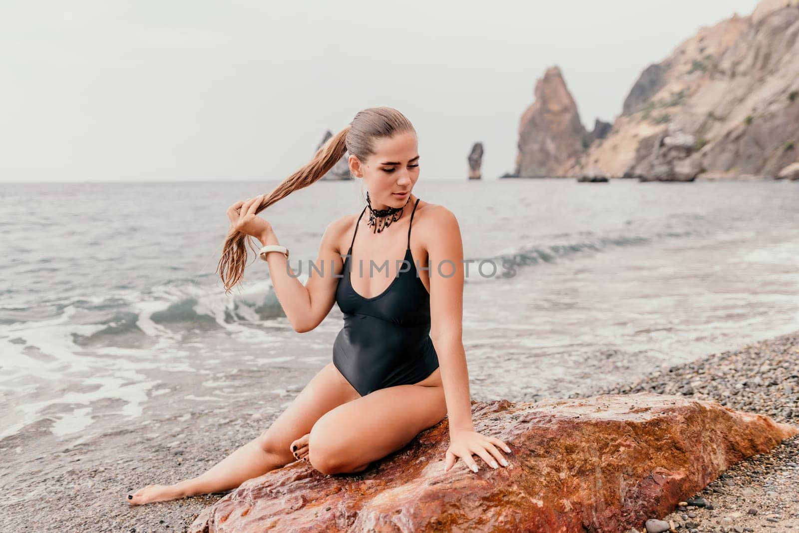 Woman travel portrait. Happy woman with long hair poses on a red volcanic rock at the beach. Close up portrait cute woman in black bikini, smiles at the camera, with the sea in the background