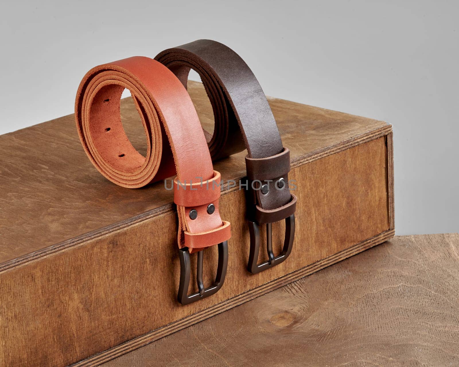 Two folded stylish leather belts with DAD embossment and metal matte buckles showcased on wooden display steps. Fashionable personalized mens accessories concept