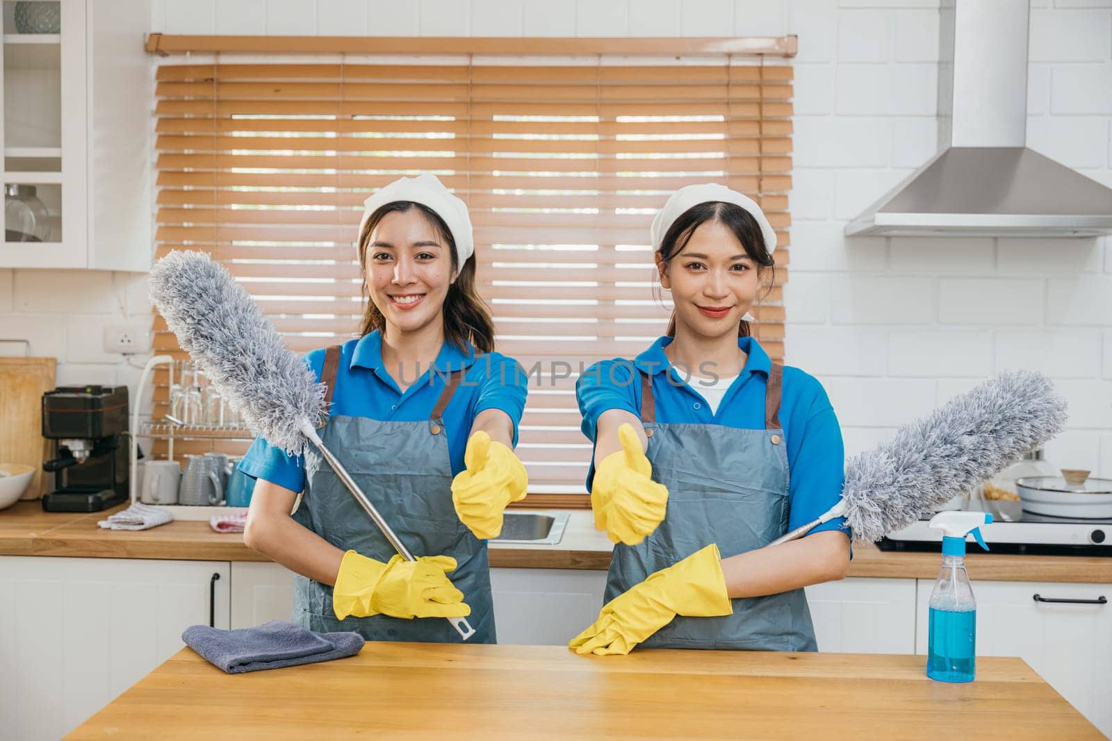 Asian cleaning team two women in uniform on kitchen counter hold duster foggy spray and rag. Depicting teamwork in efficient housework. Clean portrait two uniform maid working smiling employee. by Sorapop