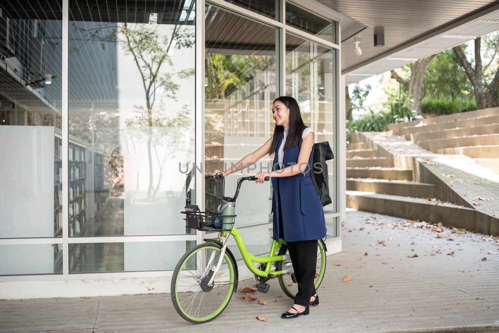 A young Asian woman commutes to work by cycling in a green city, carrying a backpack and using a reusable drinking cup to avoid harming the environment..