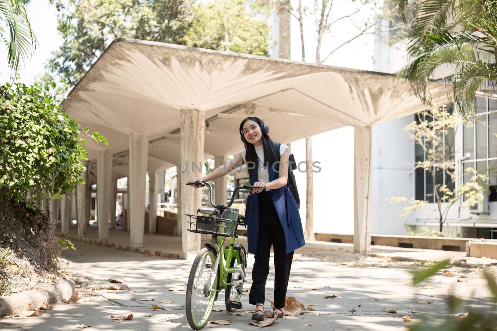 A young Asian woman commutes to work by cycling in a green city, carrying a backpack and using a reusable drinking cup to avoid harming the environment. by wichayada