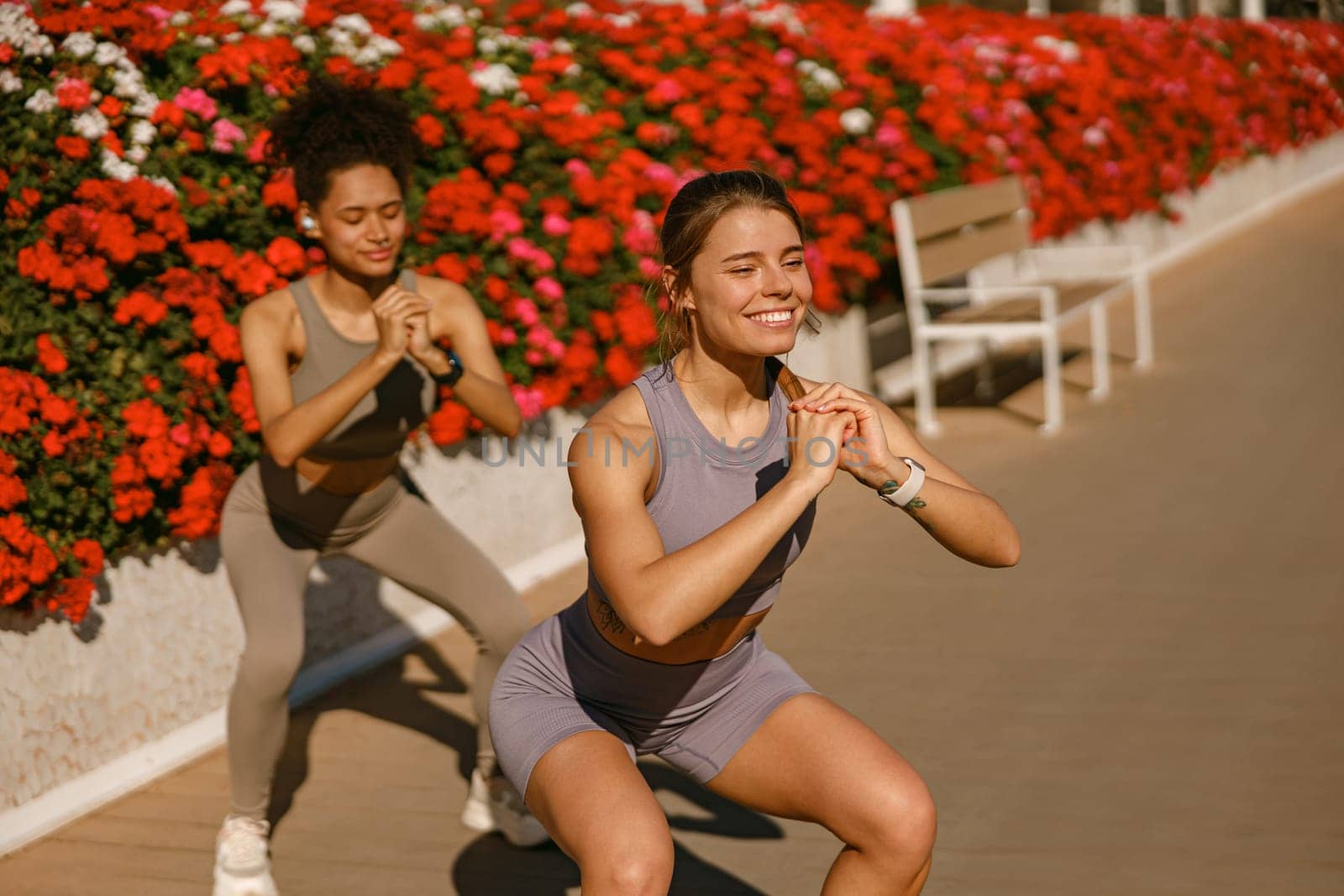 Two smiling fit women doing workout and squatting together outdoors. Active lifestyle concept