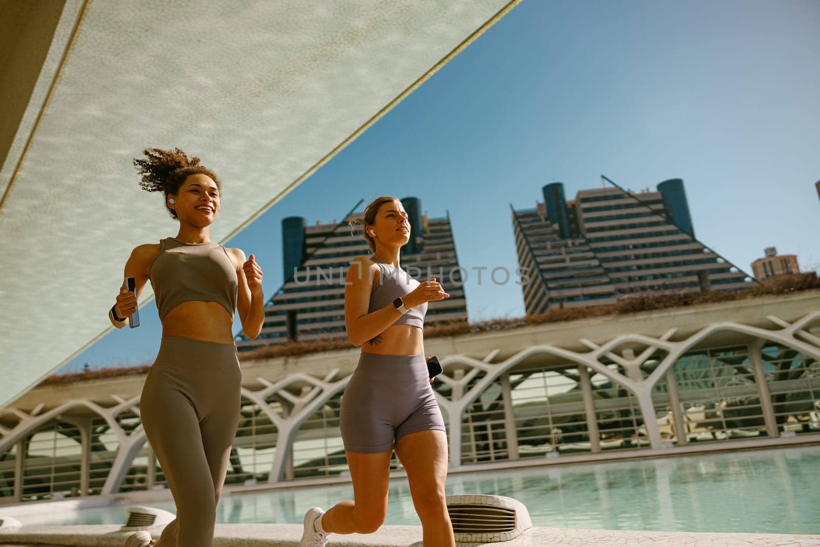 Two active women athlete running side by side along an outdoor track on modern buildings background