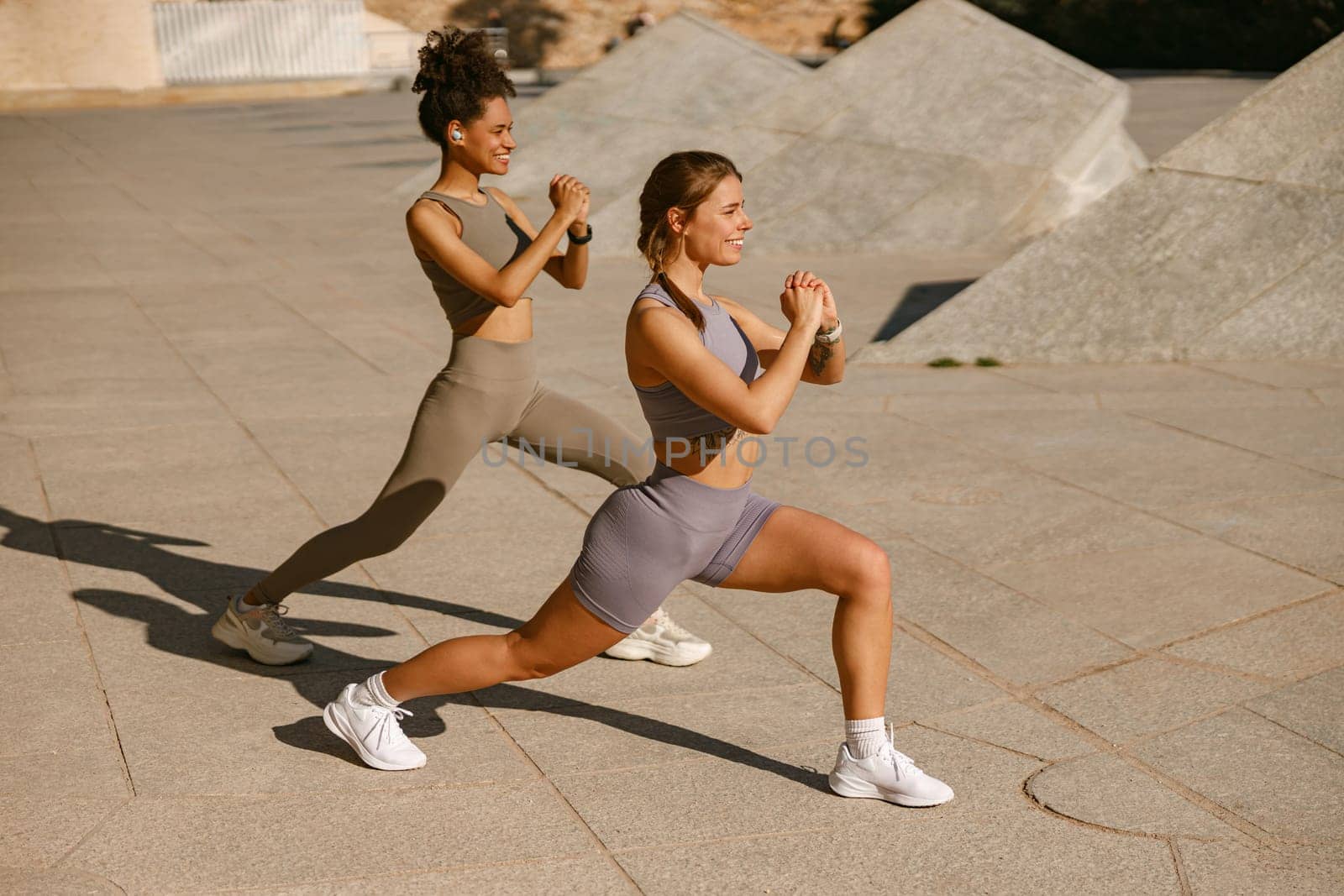 Two young sporty women in tracksuits are stretches legs outside on modern building background by Yaroslav_astakhov