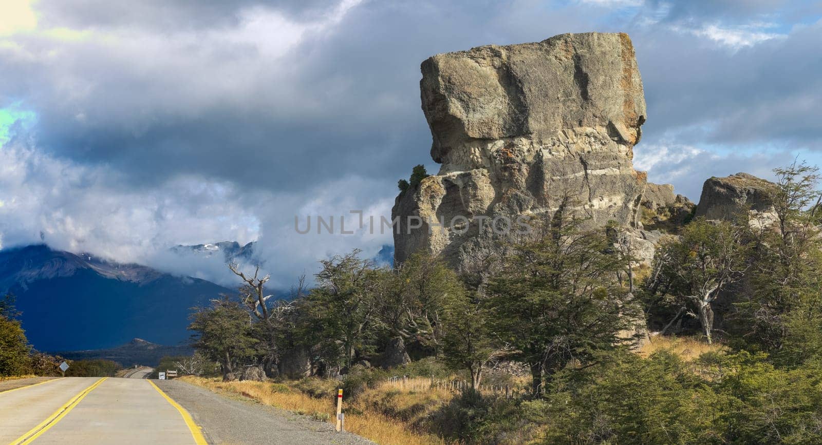 Open road with yellow stripes leads to high rocks under a cloudy sky.