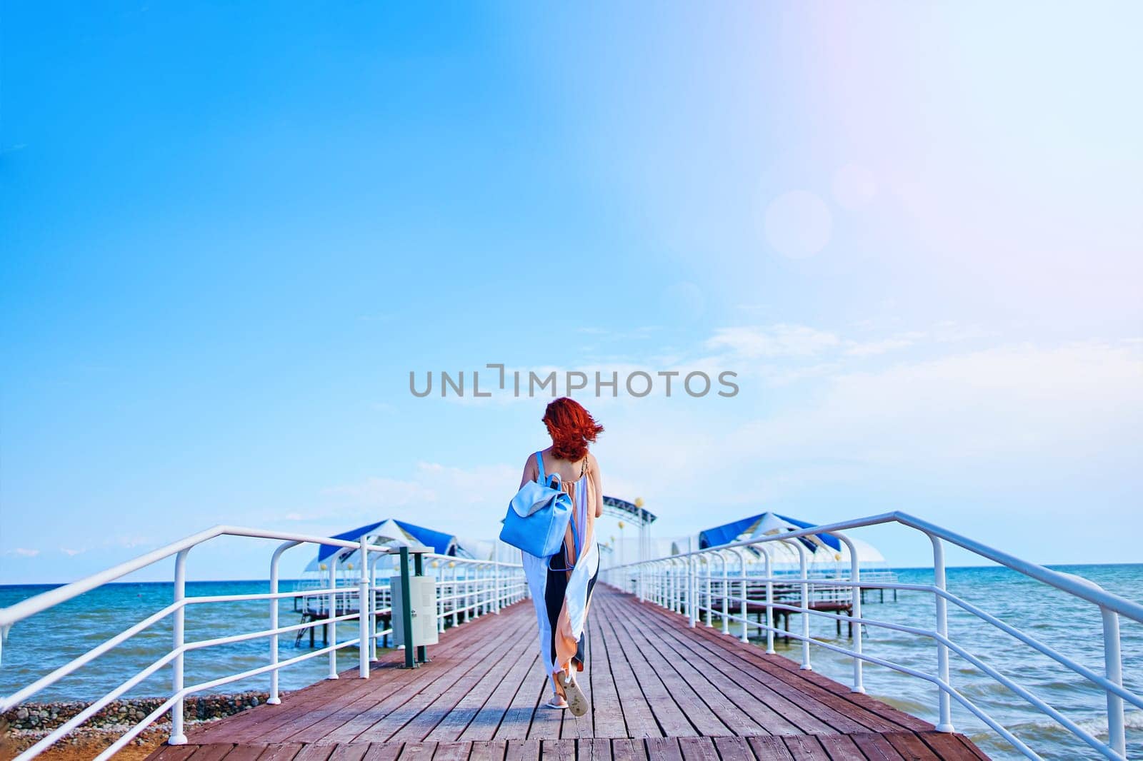 A stunning woman with fiery red hair strolls along the resort pier, adorned in a summer dress and carrying a backpack, with the sea as her picturesque backdrop.