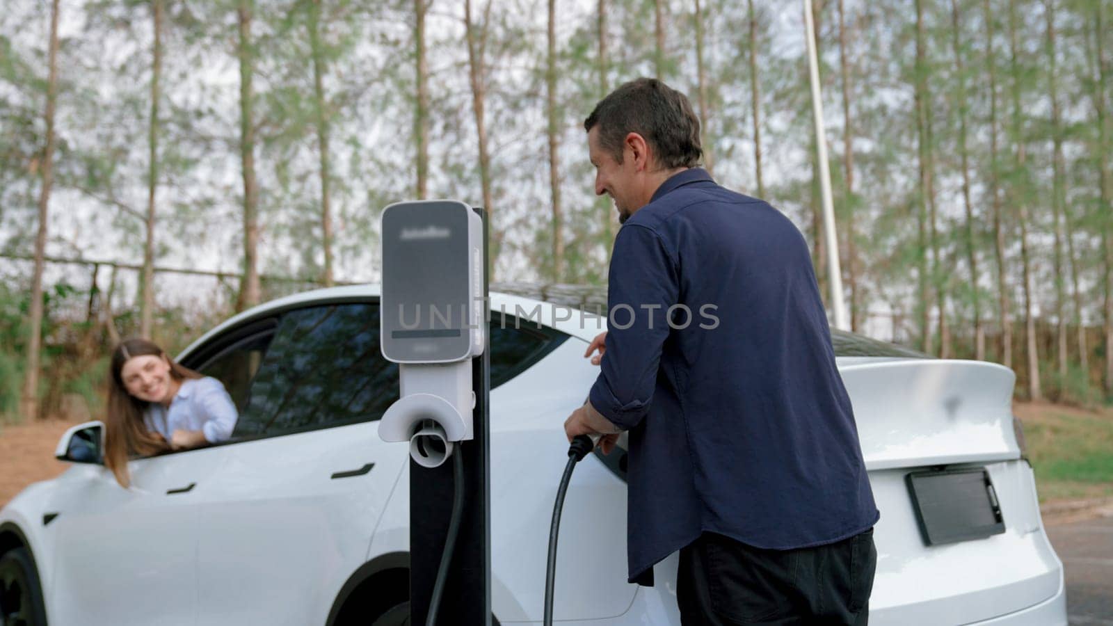 EV car recharging electricity for battery by lovey couple traveling during their road trip travel by eco friendly electric vehicle in national park autumnal forest on holiday. Exalt