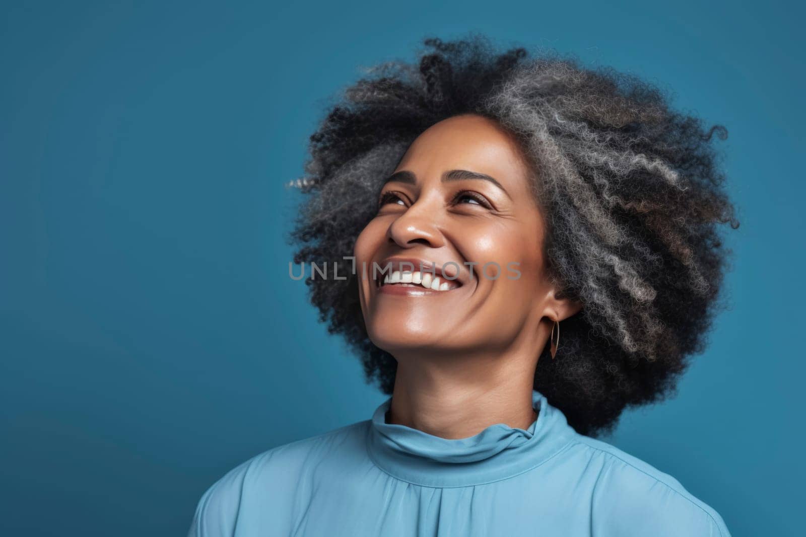 Smiling woman with curly hair in blue turtleneck on blue background. Studio portrait photography. Joy and happiness concept for design and print. Close-up with copy space