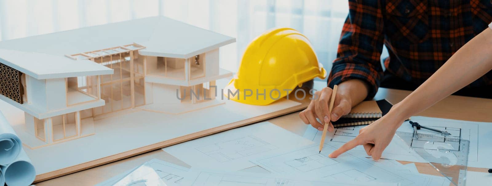 Cropped image of cooperative architect team decide and work together on meeting table with house model, safety helmet and architectural plan scatter around. Closeup. Focus on hand. Burgeoning.