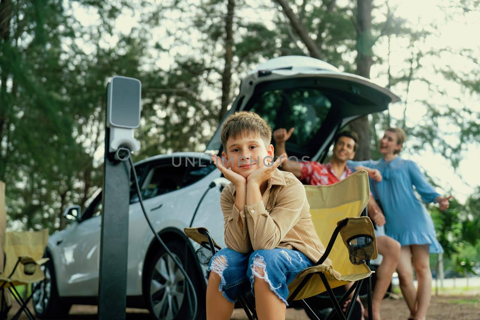 Little boy portrait sitting on camping chair with his family in background. Road trip travel with alternative energy charging station for eco-friendly car concept. Perpetual