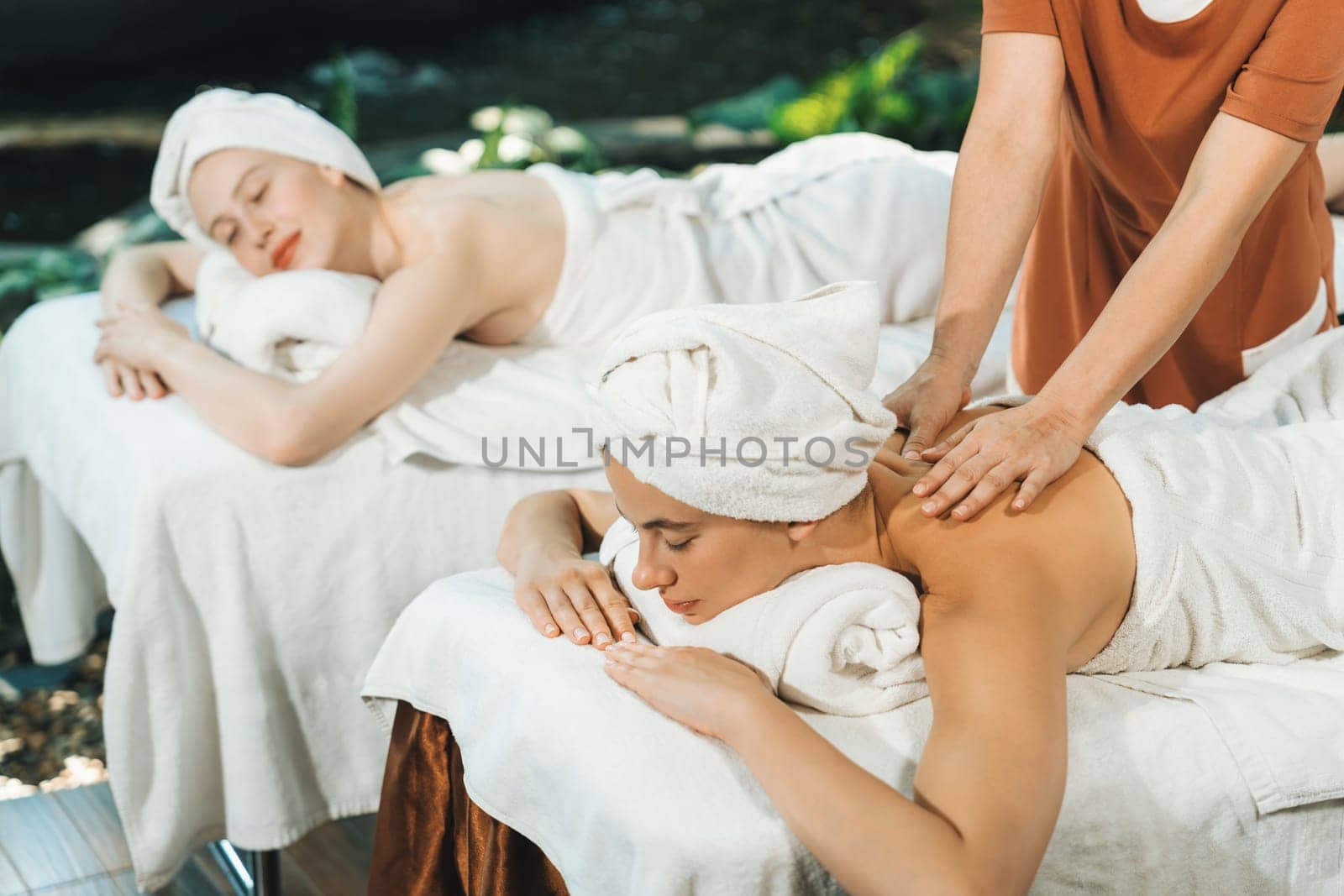 A portrait of two young attractive woman lie on bed during having back massage by a professional masseur at outdoor surrounded by peaceful natural environment. Healthy and beauty concept. Tranquility.