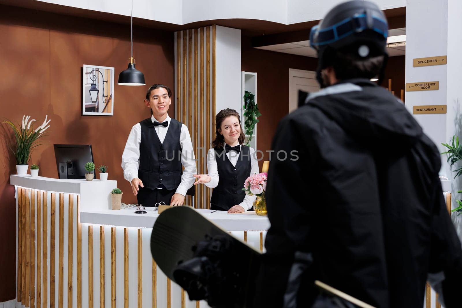 At front desk of exclusive winter resort, two receptionists warmly welcoming male guest in snow gear. Tourist wearing winter jacket and carrying snowboarding equipment arrives in hotel lobby.