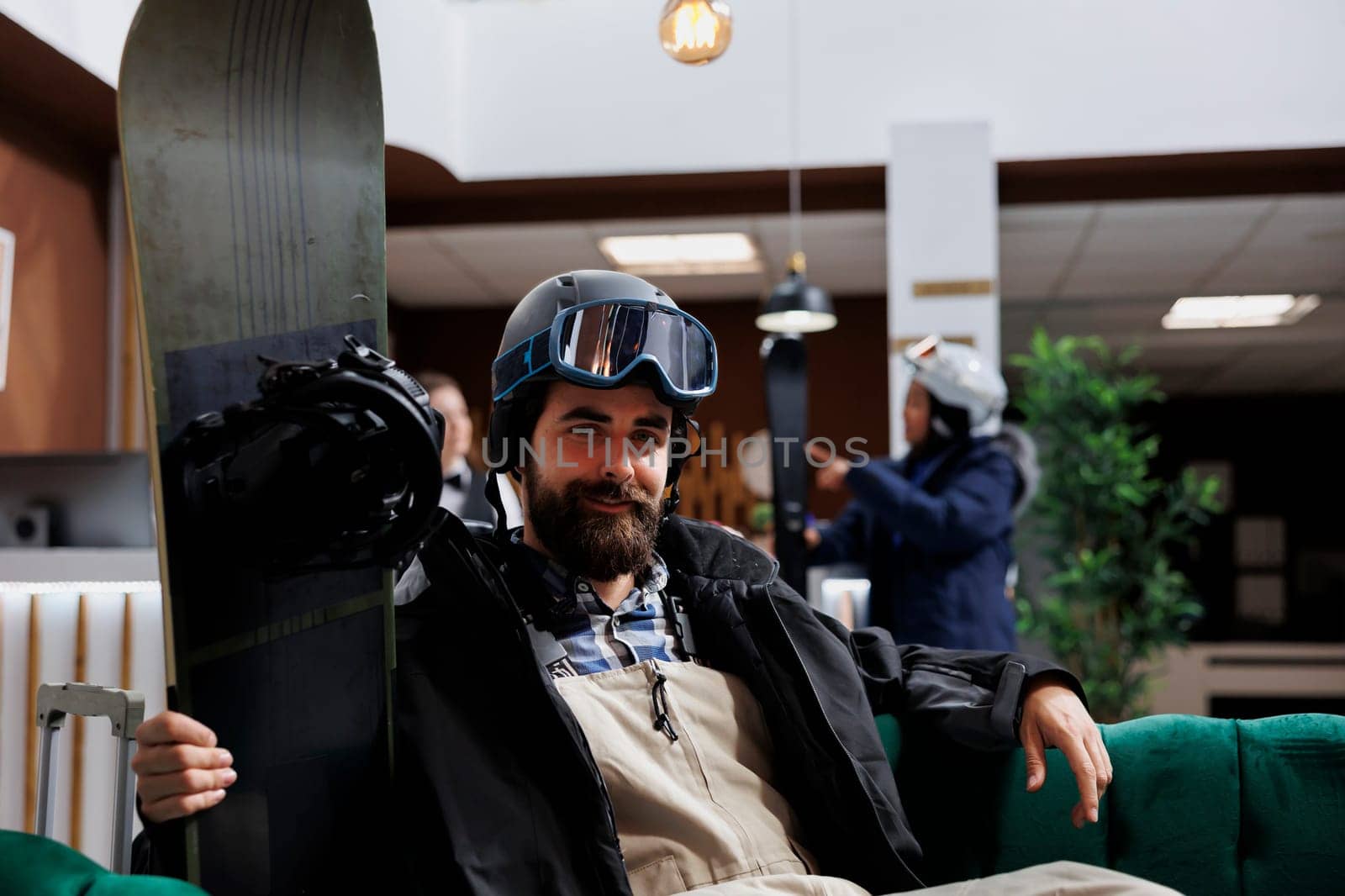 Portrait shot of caucasian male tourist wearing snow gear and holding snowboard in hotel lobby. Image showing man with winter clothing sitting in ski resort lounge with snowboarding equipment nearby.