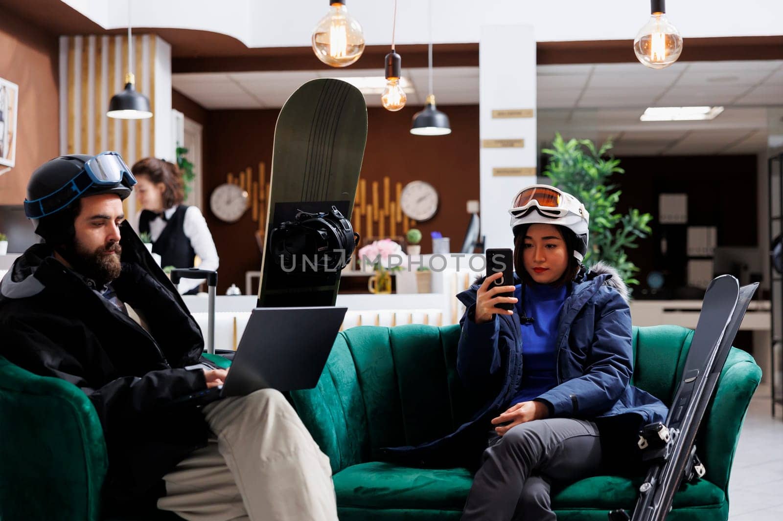 Young couple enjoys winter vacation by lounging in the ski resort lounge area with their digital gadgets. A man with a laptop and a lady with smartphone take a break from skiing and snowboarding.