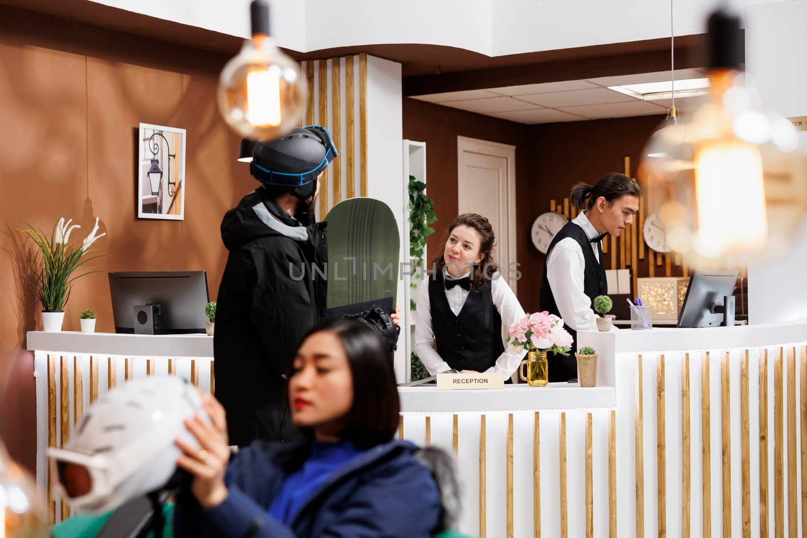 Young man wearing helmet and holding snowboard arrives at hotel reception greeted by friendly staff. Guests in snow gear going for wintersport activity at luxury ski mountain resort.