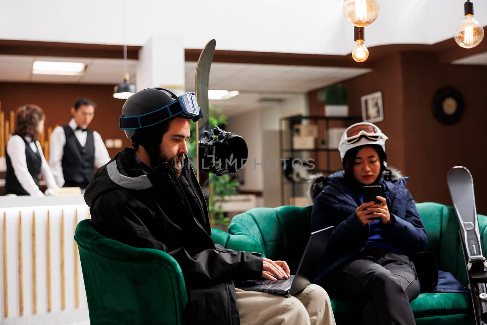 Young couple relaxing in ski resort lounge area with their digital devices enjoying winter holiday. Man with laptop and woman with cell phone taking break from skiing and snowboarding activities.