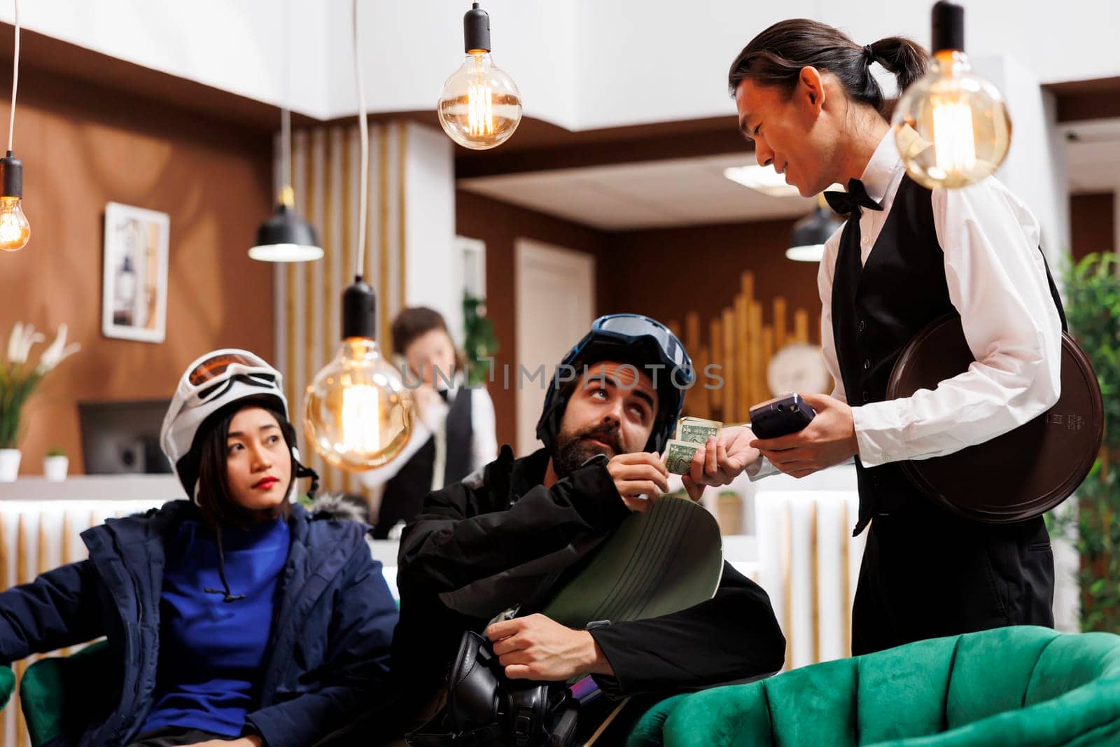 Friendly asian waiter receiving money tip from couple with snow clothing in lounge area of ski mountain resort. Healthy boyfriend and girlfriend thanking employee for good service in winter vacation.