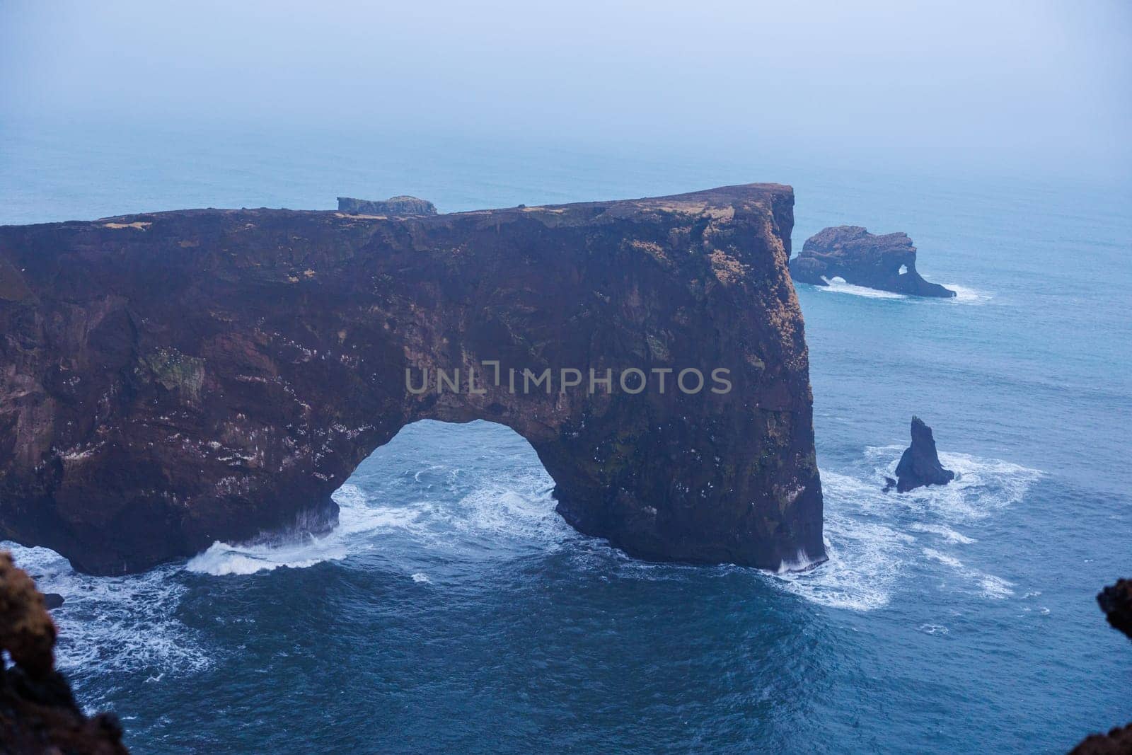 Stone coastline arch on dyrholaey peninsula with nordic foggy landscape and rocky cliffs. Spectacular shoreline stoney gate in iceland, beautiful wilderness with harbor bay view.