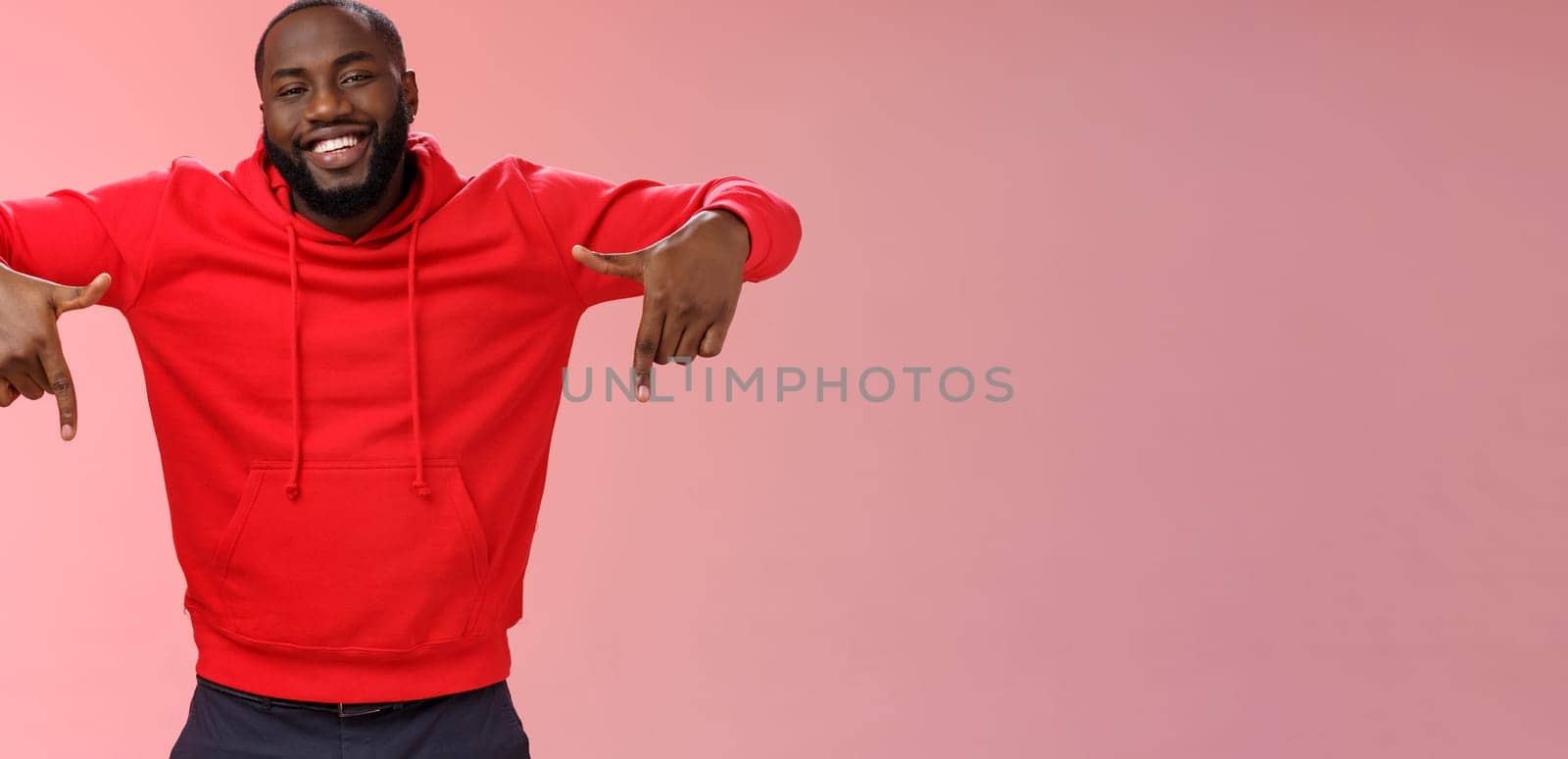 Happy charismatic smiling male african american entrepreneur proudly pointing down presenting his own company product gladly showing accomplishment indicating downwards, standing pink background.