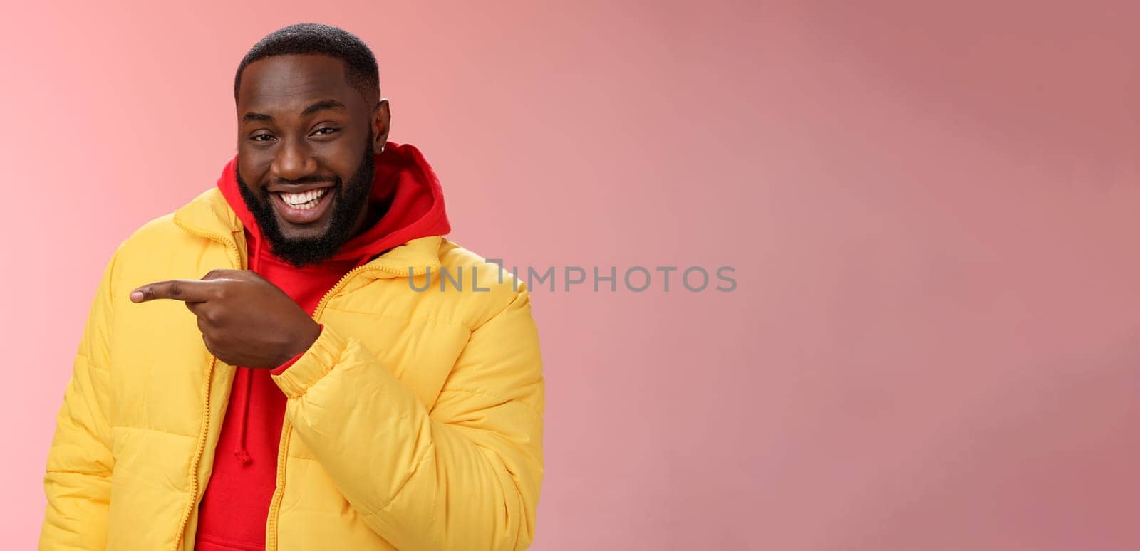 Lifestyle. Charismatic carefree handsome black bearded guy in yellow jacket laughing friendly look camera chat pointing left show cool place hang out standing joyful pink background talking having fun.