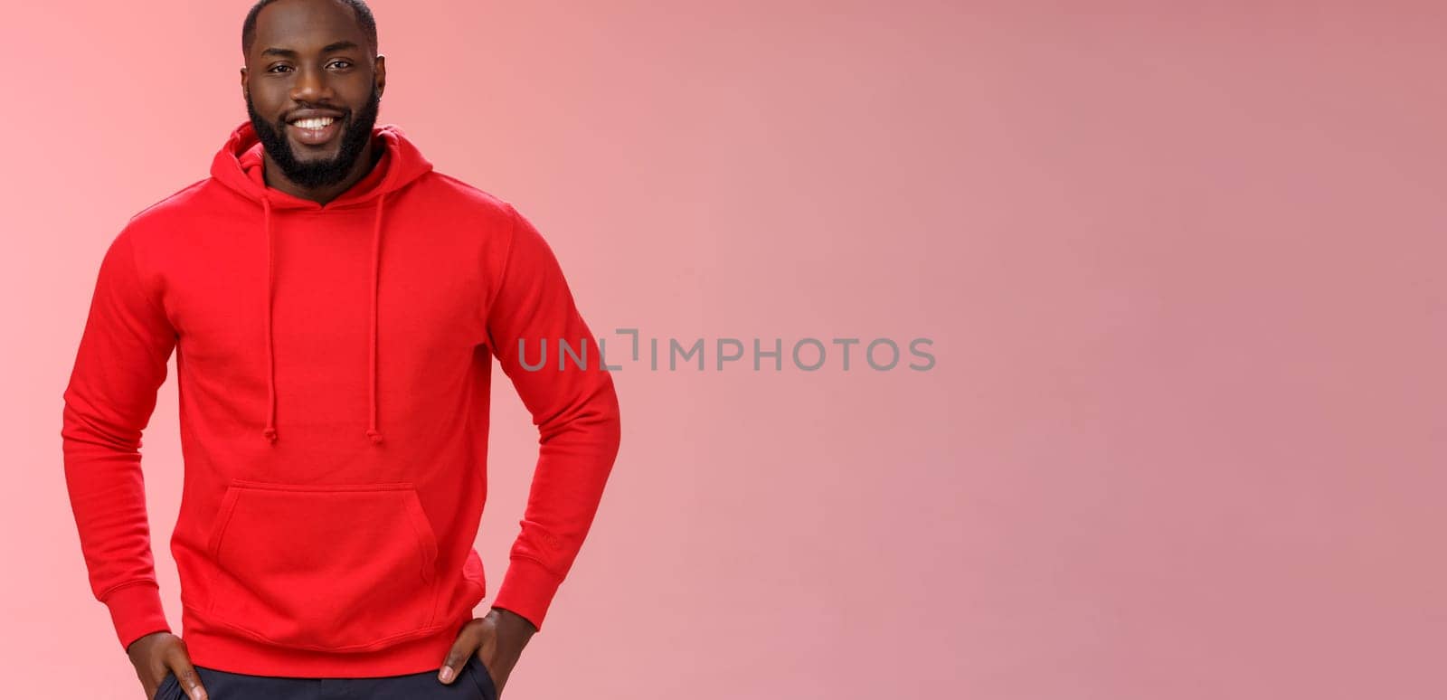 Handsome young happy black bearded guy white perfect smile hold hands pockets casually standing pink background talking have interesting nice conversation, wearing red hoodie. Copy space