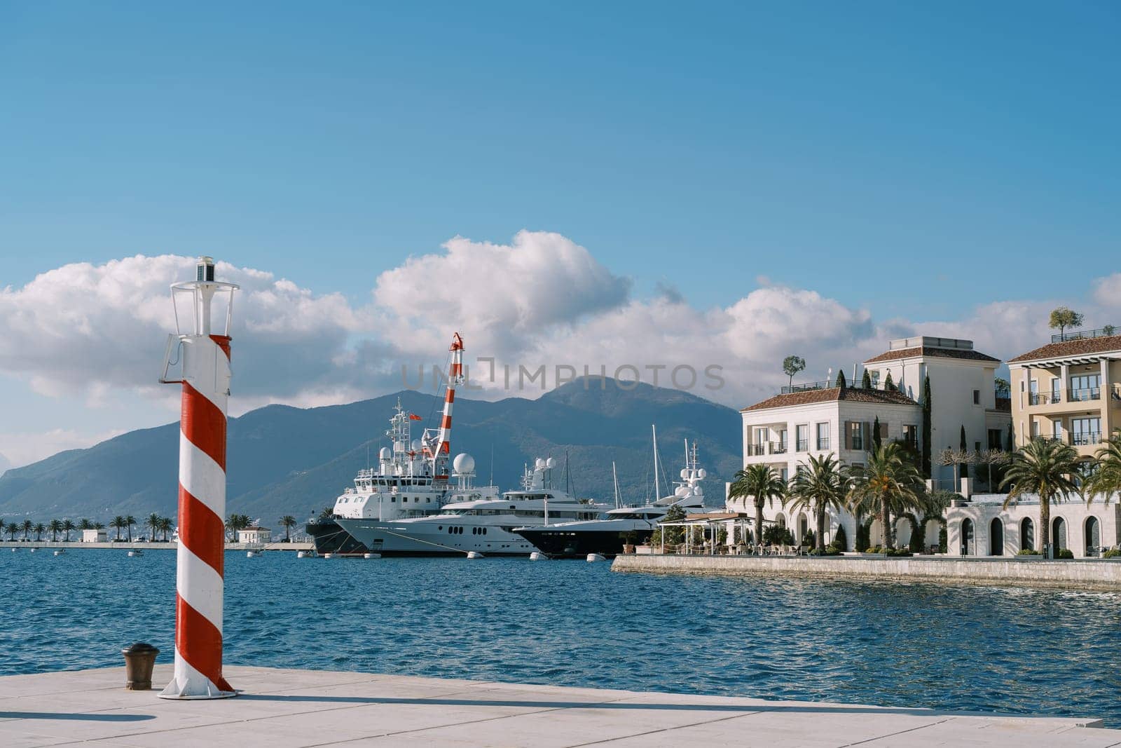 Small striped lighthouse stands on the pier of a luxury marina with moored yachts by Nadtochiy