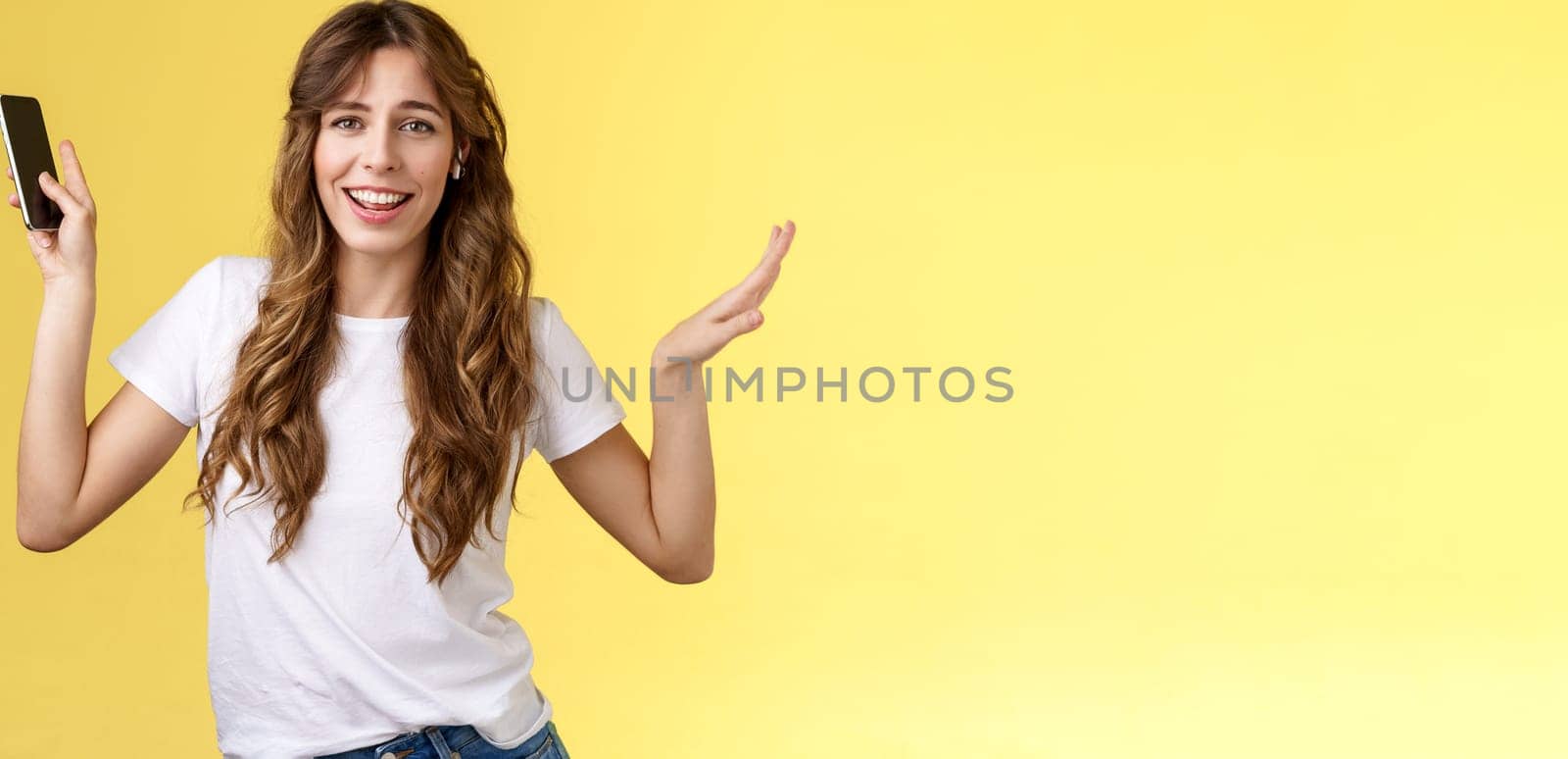 Carefree good-looking lively sociable curly-haired girl having fun wearing wireless earbuds dancing joyfully listening music moving rhythm song hold smartphone raising hands up smiling camera amused. Lifestyle.