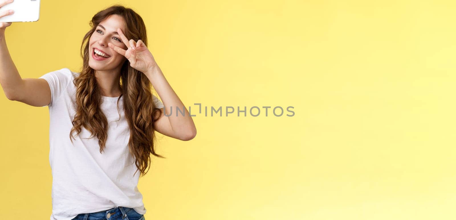 Joyful charismatic good-looking curly-haired woman white t-shirt show peace victory sign smiling broadly posing tender friendly expression smartphone front camera take selfie yellow background by Benzoix